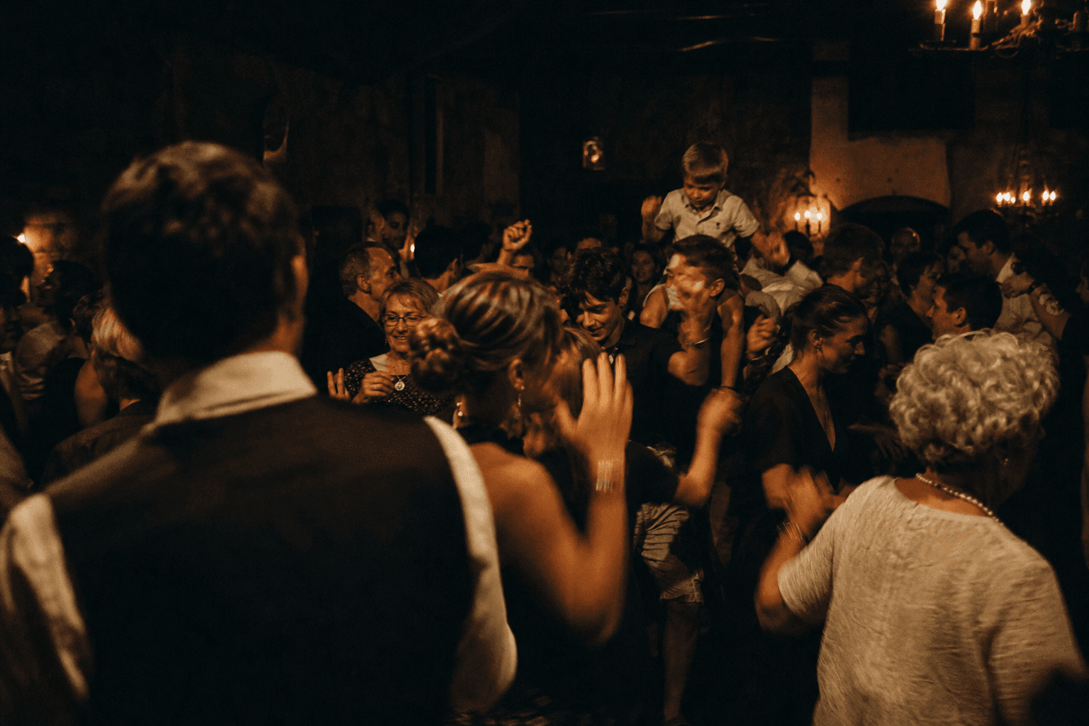 Une piste de danse pleine à 23:00, trois générations qui dansent ensemble sous une lumière de bougies.